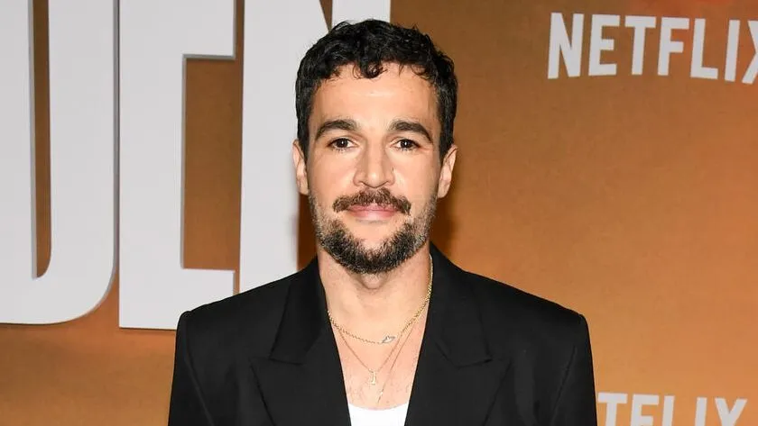 A bearded man in a black blazer stands against an orange backdrop with white Netflix lettering.