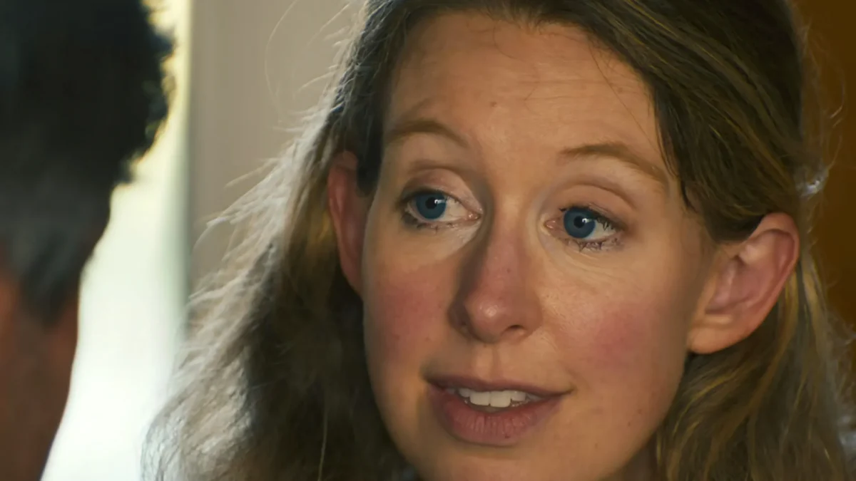 A close-up shows a blue-eyed woman speaking to a blurred person in the foreground.