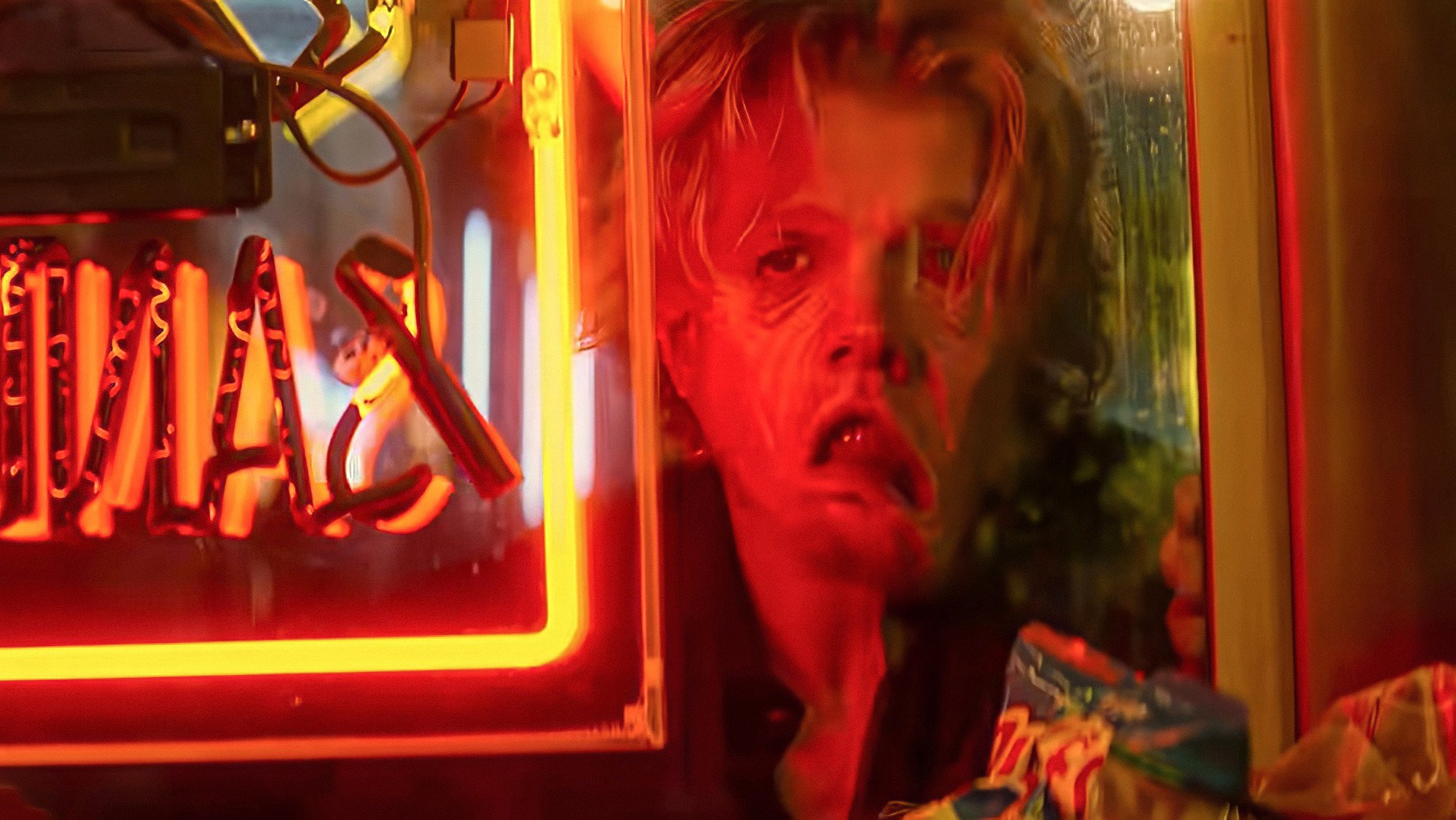 A person peers through a rain-streaked window beside a glowing red neon sign.