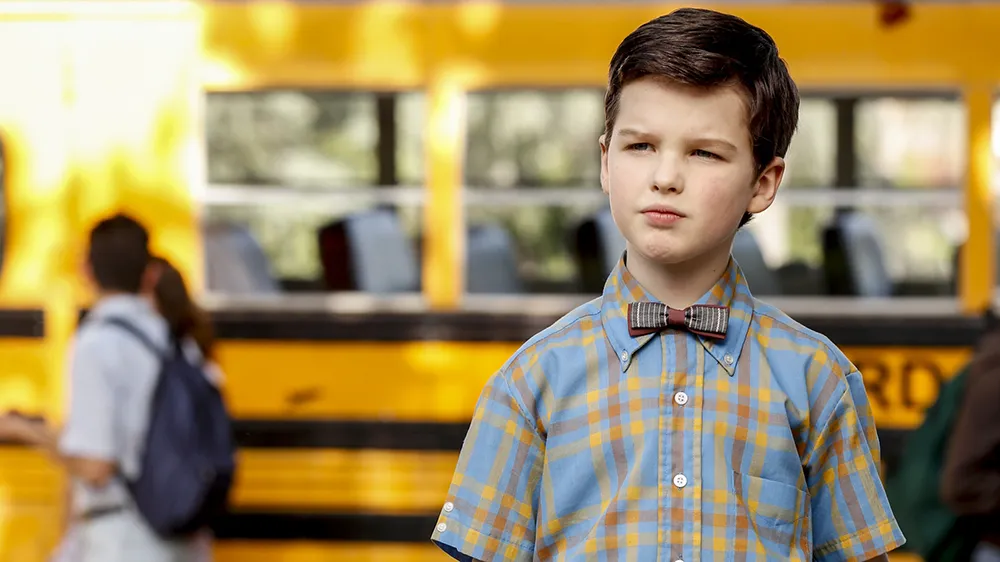 A young boy wearing a plaid shirt and bow tie stands in front of a yellow school bus.
