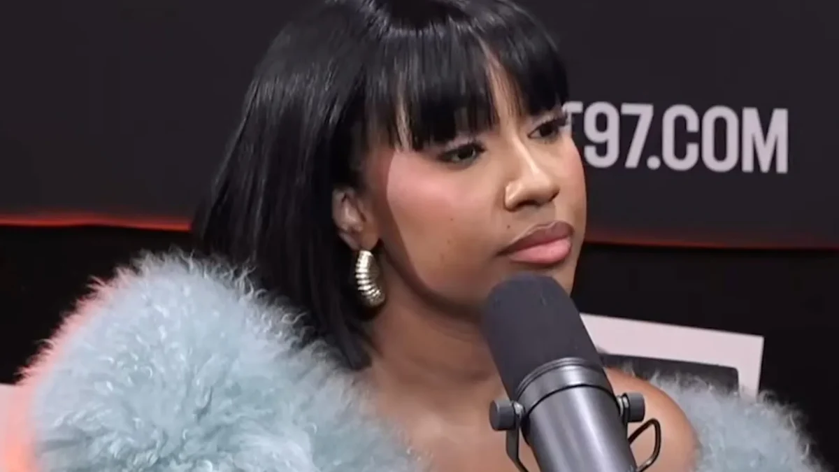 Woman in a blue fur jacket sits behind a microphone against a dark studio backdrop.