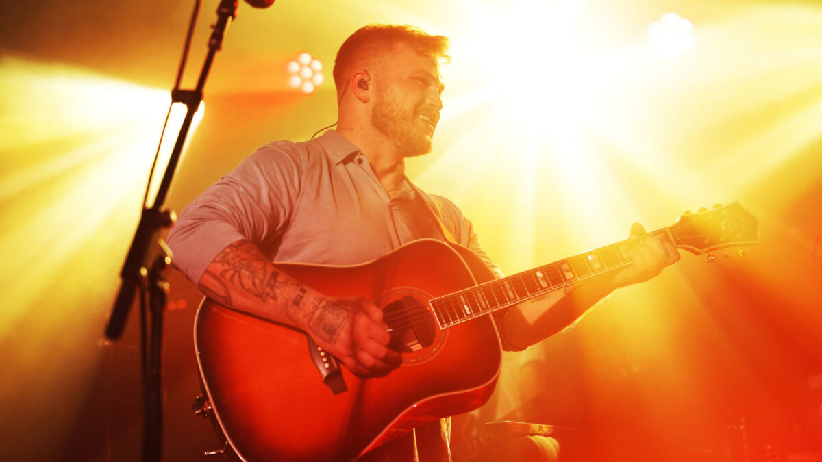 A bearded man plays an acoustic guitar under bright orange stage lights.