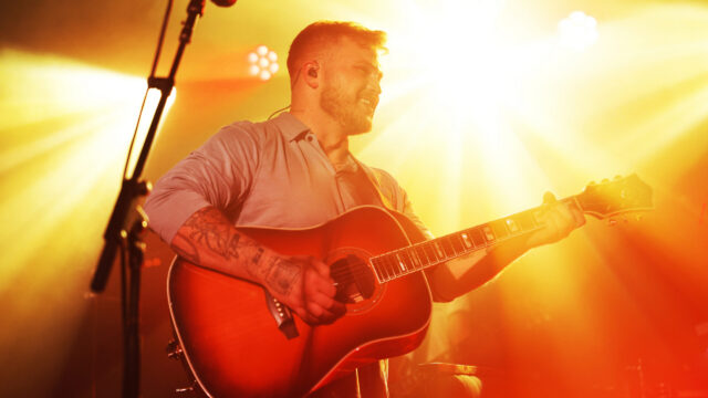 A bearded man plays an acoustic guitar under bright orange stage lights.