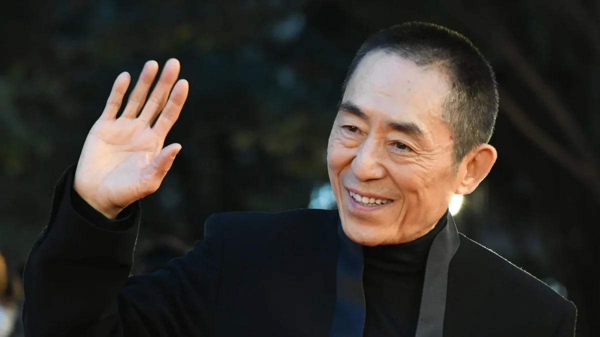 A smiling man in a dark jacket raises his hand in a wave.